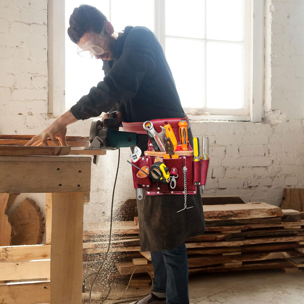 A carpenter wearing goggles and Fueri maroon leather toll pouch full of all tools, standing by a workbench, wood pieces in background.